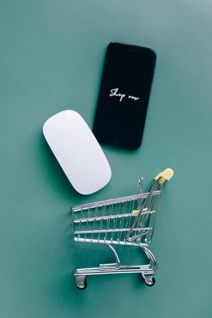 Flat lay of a miniature shopping cart, smartphone, and computer mouse on teal background.