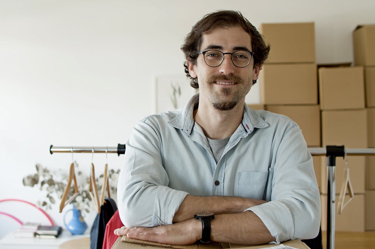 Young businessman with glasses smiling in a startup setting with boxes behind him.