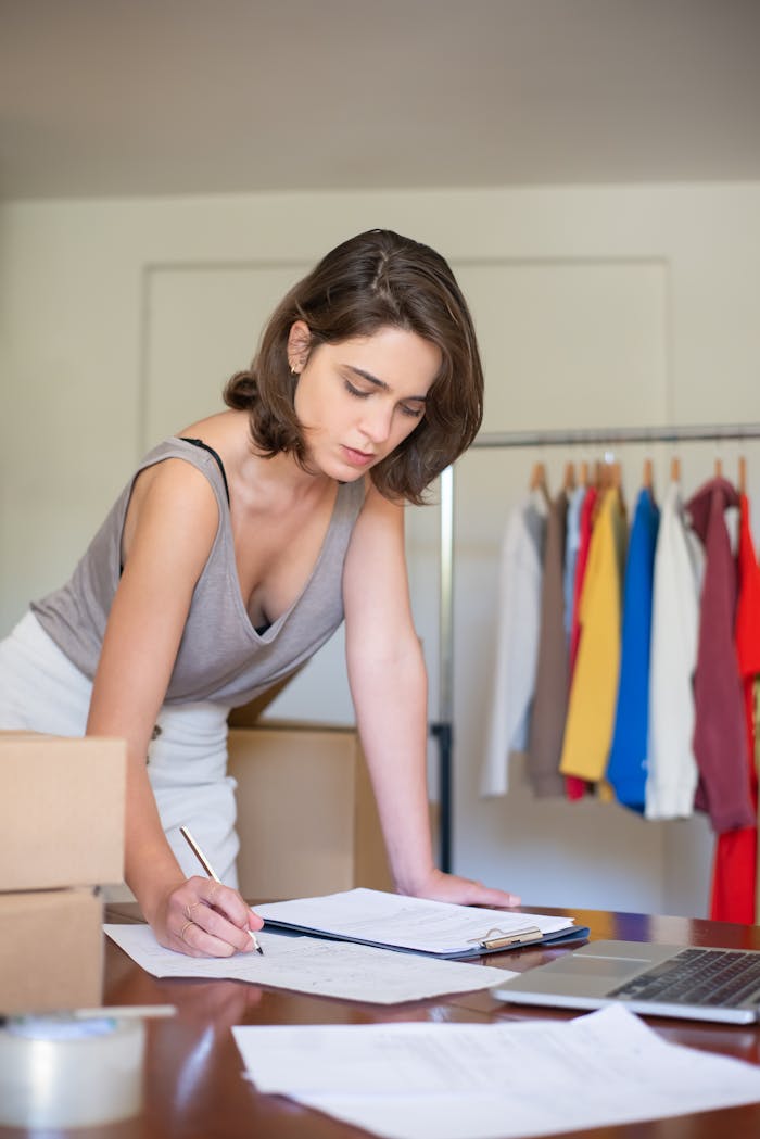 A young woman writes in her online store, surrounded by clothes and shipping boxes.